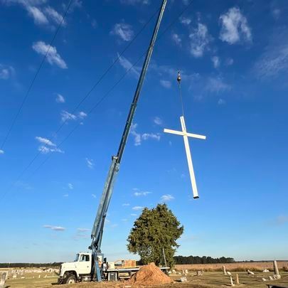 A large white cross is being lifted by a crane in a cemetery.