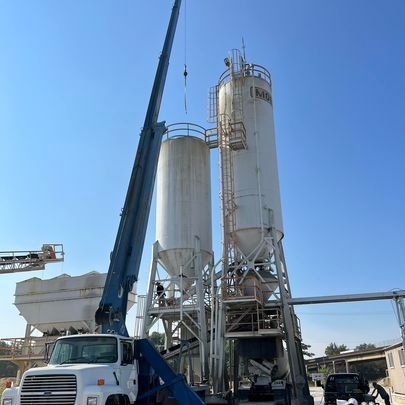 A truck with a crane attached to it is parked in front of a concrete plant.