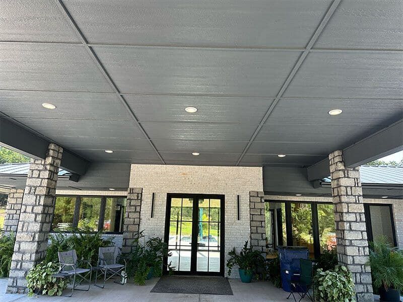 The entrance to a building with a gray, paneled ceiling, stone pillars, double glass doors, and potted green plants.
