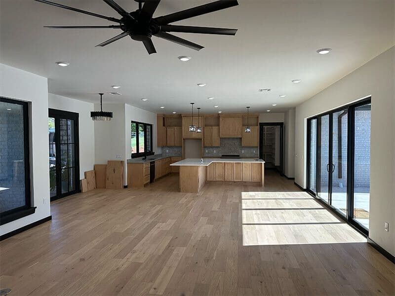 A spacious, modern open-plan kitchen and living area with light wood floors, natural wood cabinets, and large glass doors.