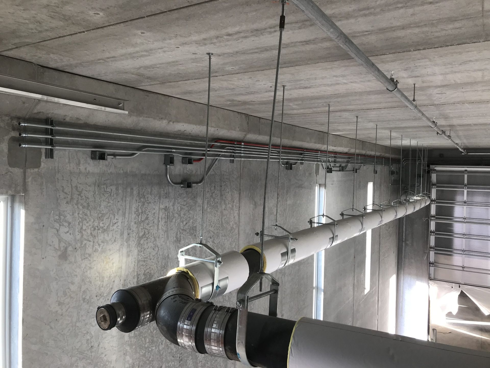 Pipes and electrical conduits suspended from a concrete ceiling in a commercial or industrial space.