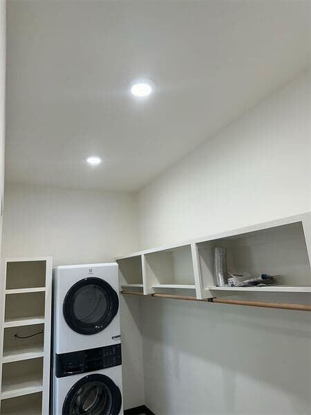 A white laundry room features a stacked washer and dryer unit, a tall shelving unit, and wall-mounted storage shelves.