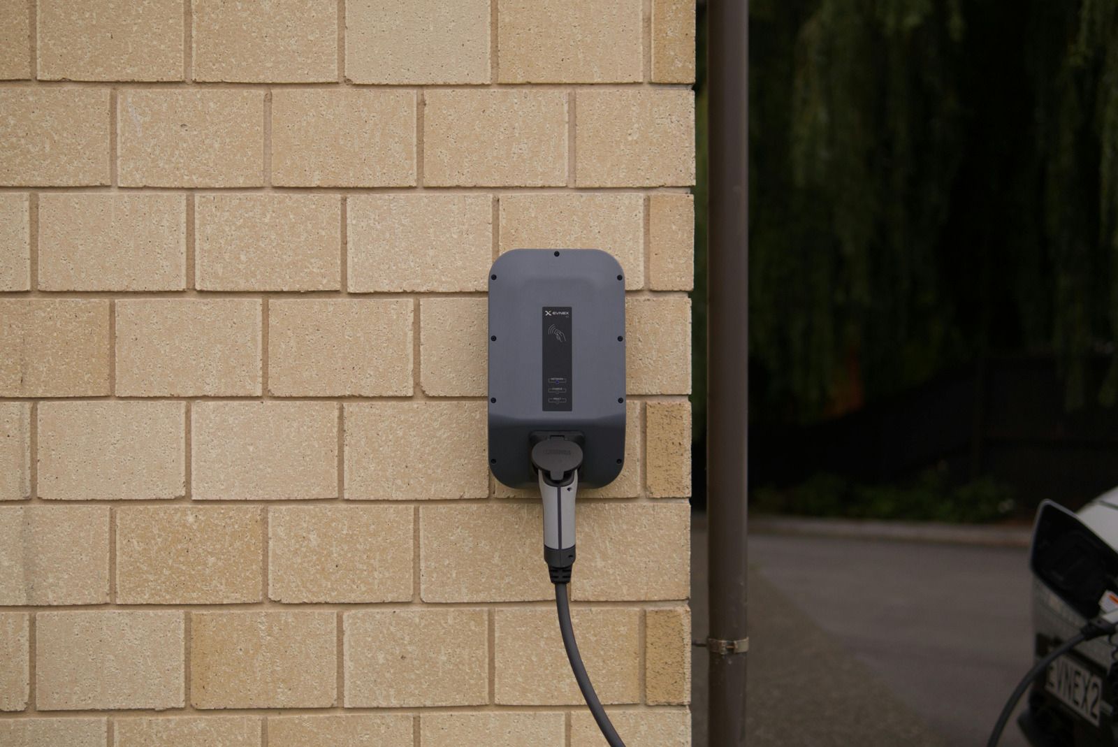 A grey electric vehicle charging station mounted on an exterior light-colored brick wall with a charging cable plugged in.