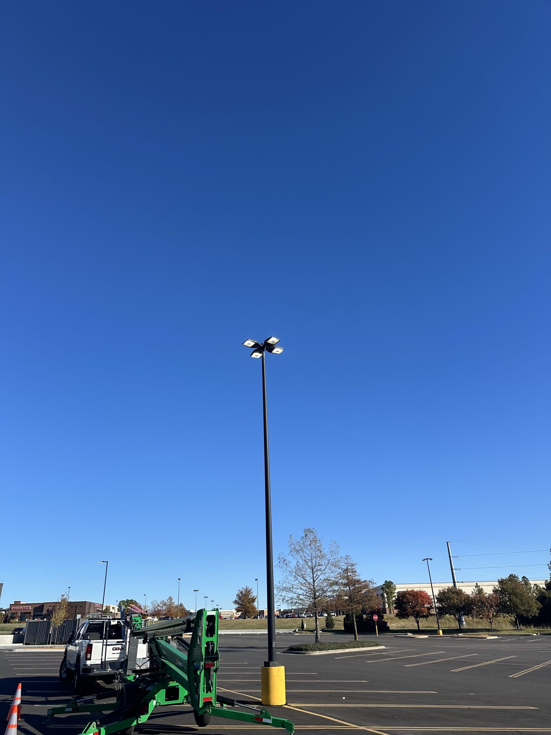 A tall parking lot light pole with four lights stands against a clear blue sky, with a green lift vehicle parked nearby.