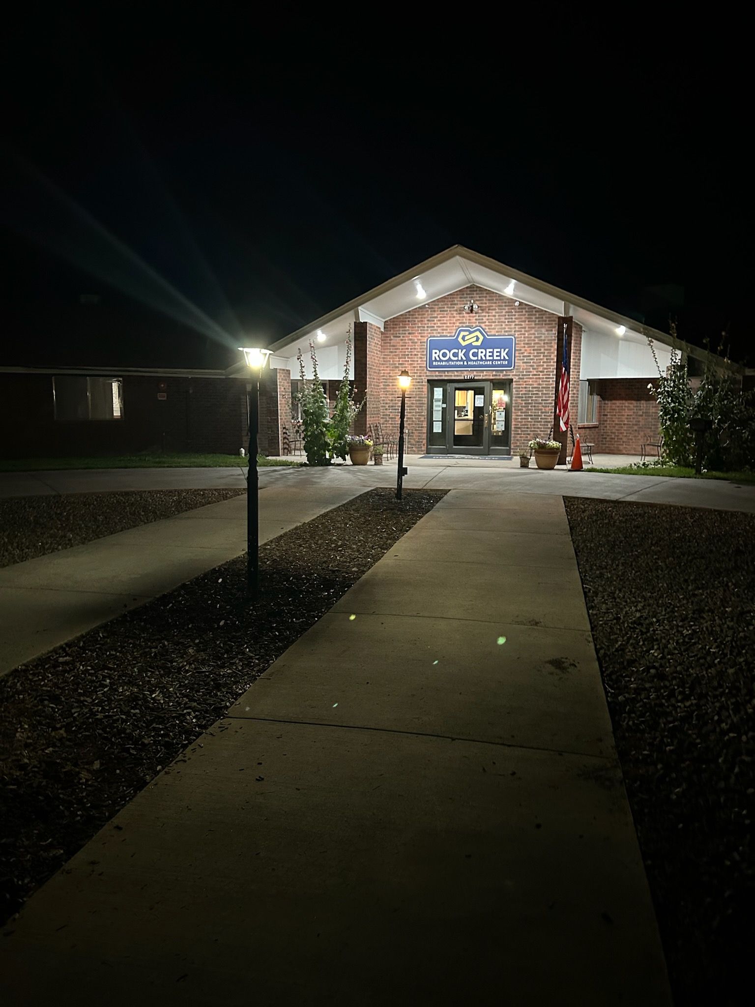 A brick building entrance illuminated at night, viewed from a path leading up to the center door under warm outdoor lights.