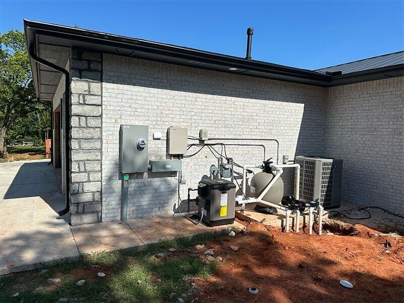 Outdoor pool equipment and electrical boxes mounted on a light brick house wall next to an area of exposed dirt.