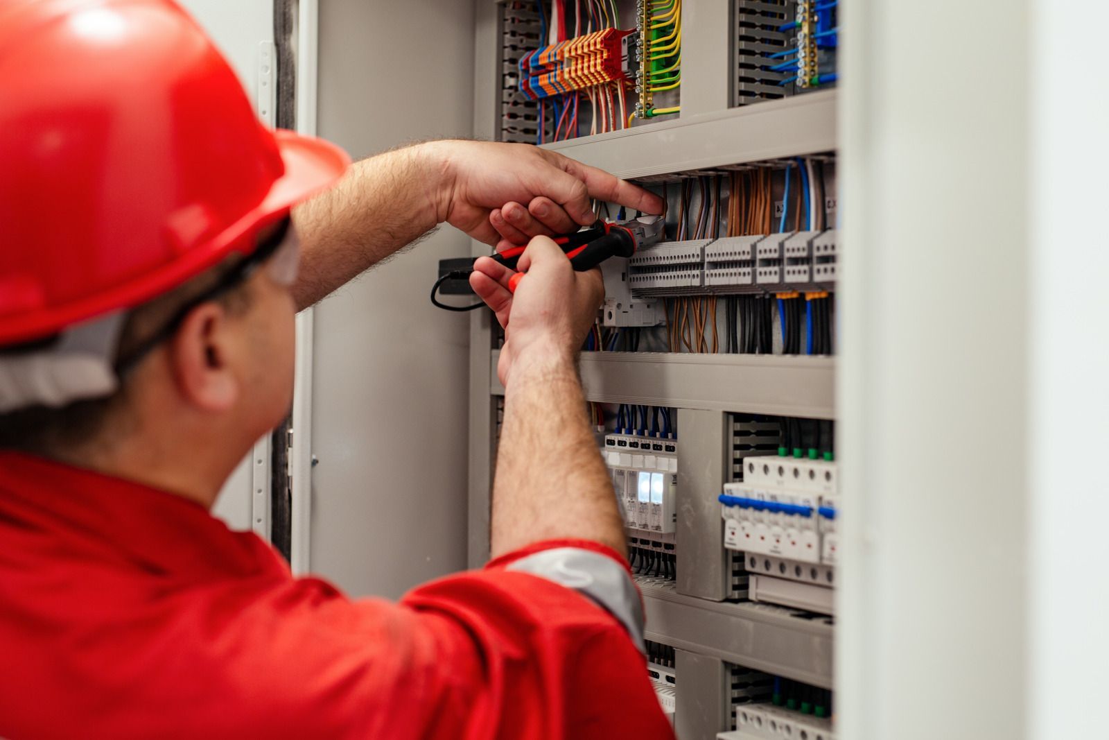 A worker in a red uniform and hard hat uses a multimeter to inspect electrical wiring in a control panel.