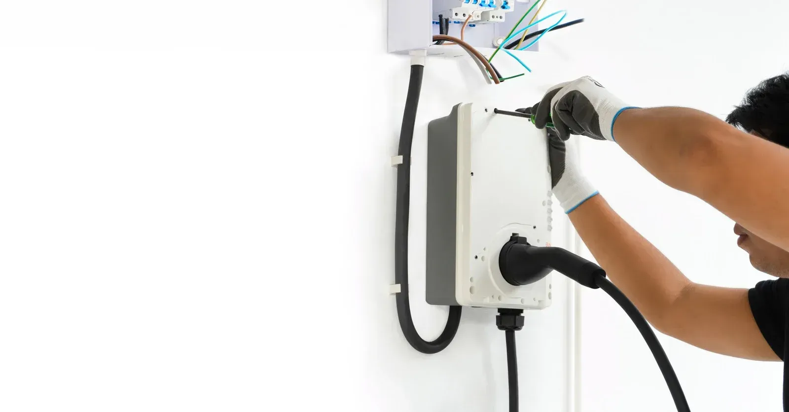 A person in work gloves installing a white electric vehicle charging station on a plain white wall.