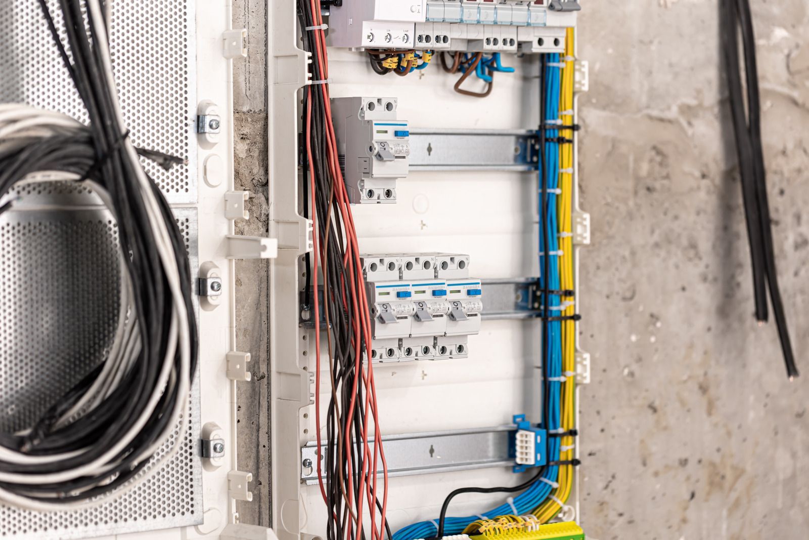 An open electrical distribution board with mounted circuit breakers, wiring, and cable management on a concrete wall.