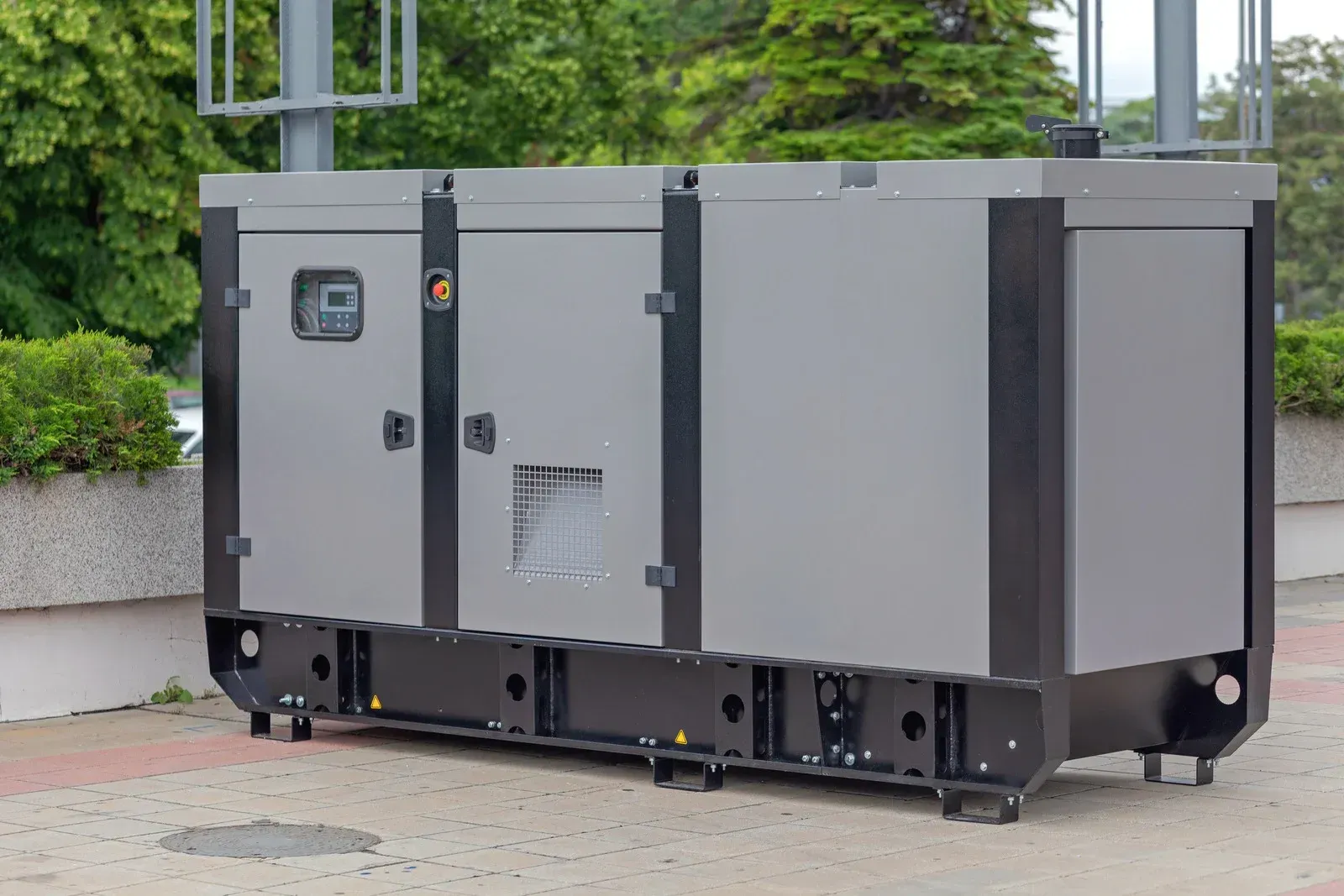 A gray industrial generator with a black base parked outdoors on a paved surface near greenery.