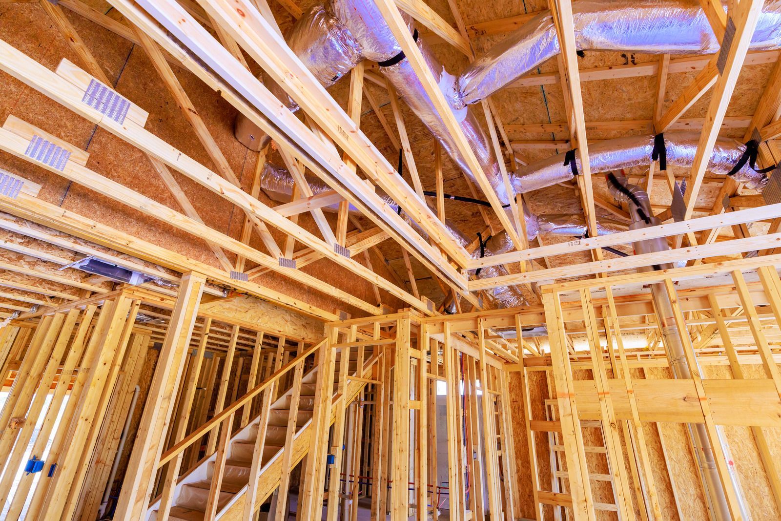Interior view of a house under construction with exposed wooden framing, roof trusses, and installed ventilation ducts.