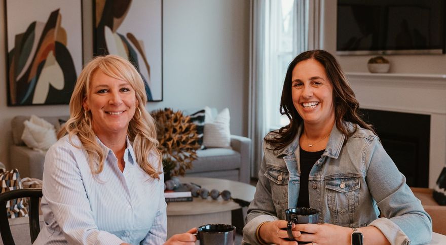 Two people sitting at a table with mugs in a living room with abstract art, a sofa, and a fireplace in the background.