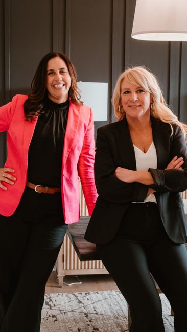 Two smiling professionals stand in an office; one wears a bright pink blazer and black top, the other a black blazer.