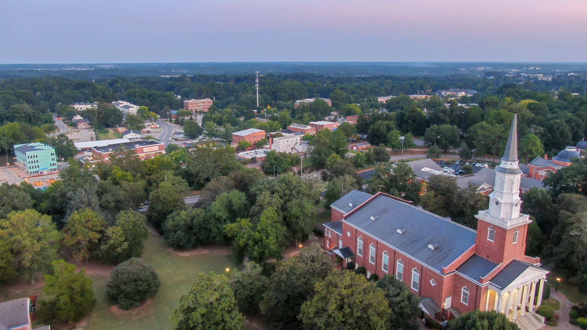 An aerial view of a red brick church with a tall steeple, surrounded by lush trees, overlooking a small town at dusk.