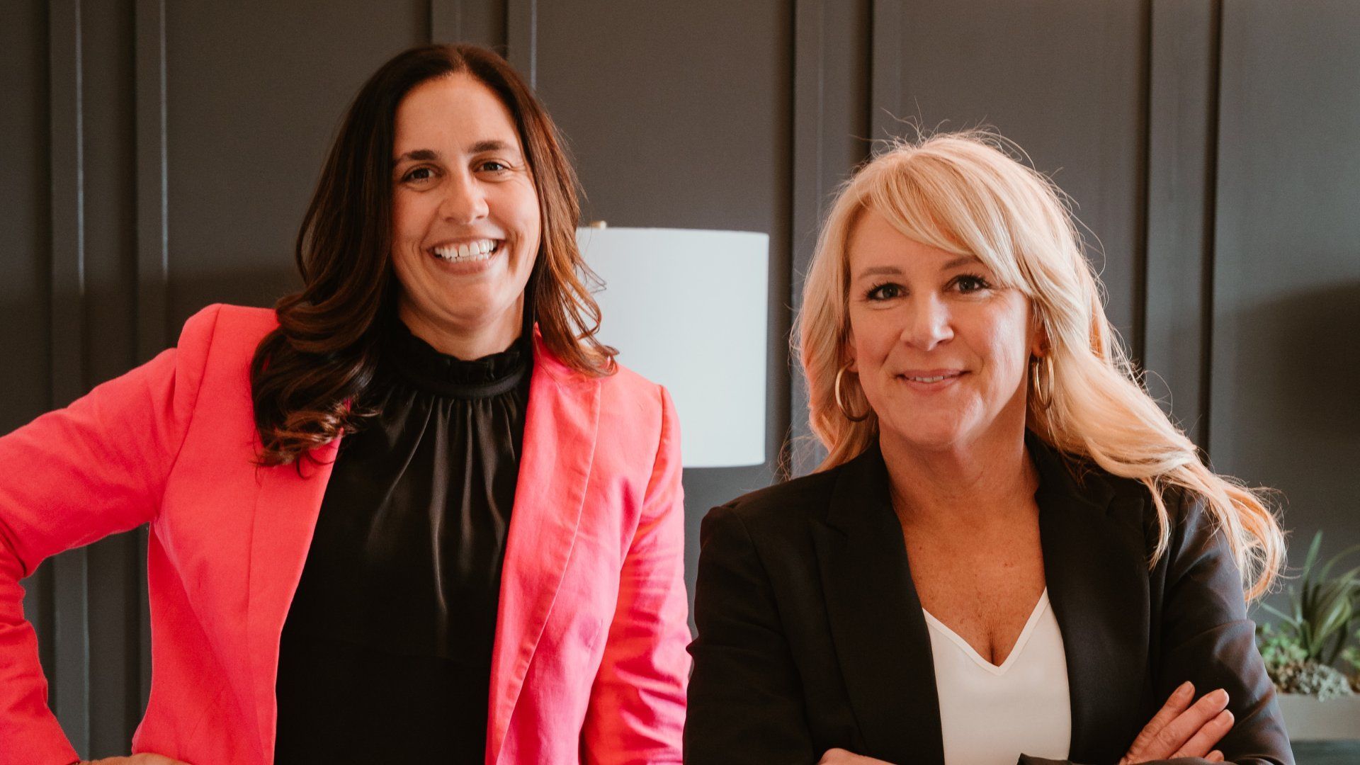 Two women smiling in a professional setting; one in a pink blazer, the other in a black blazer with crossed arms.