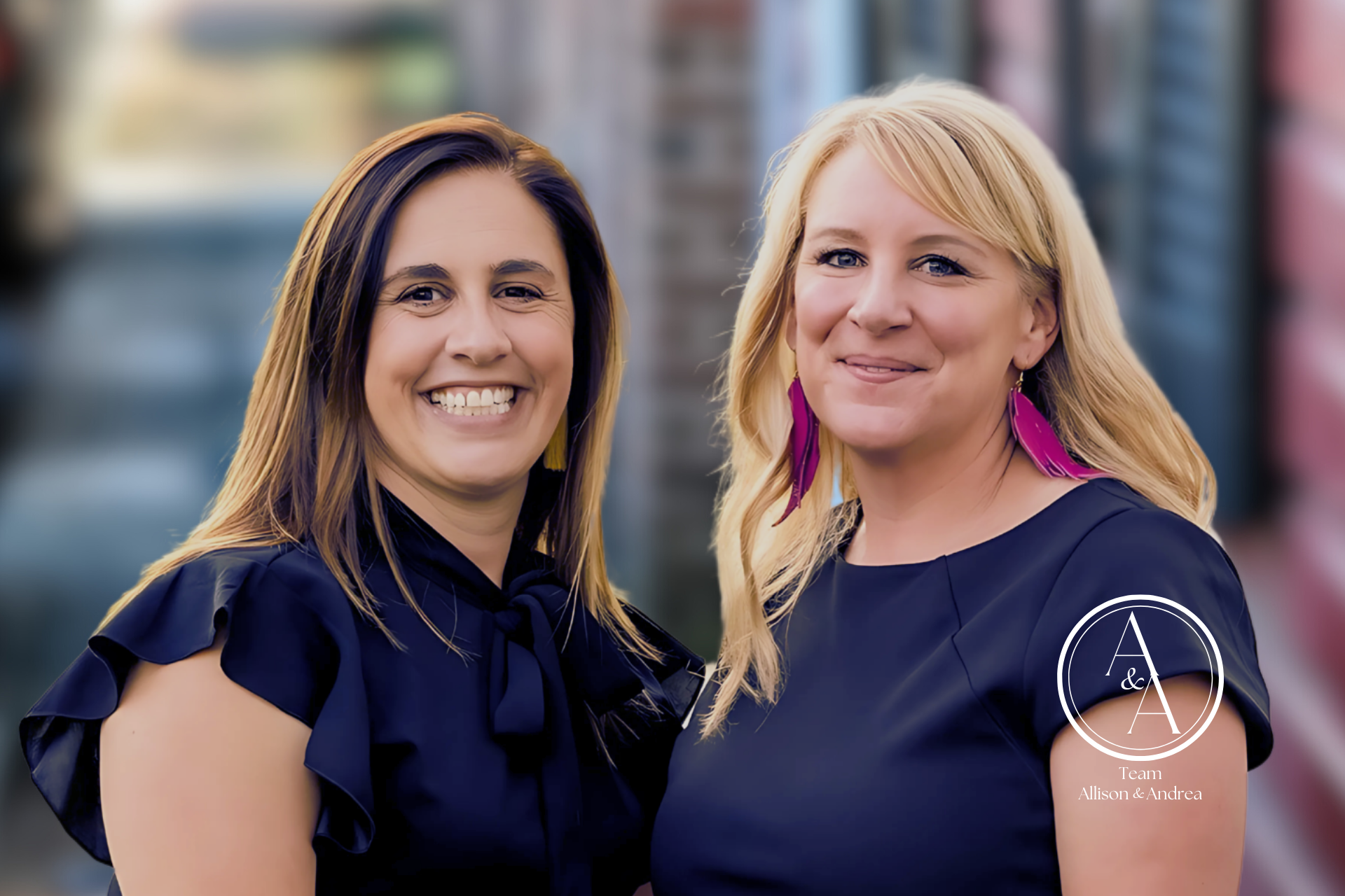 Two women smiling, posing for a photo. One wears a dark blouse, the other a dress and pink earrings.