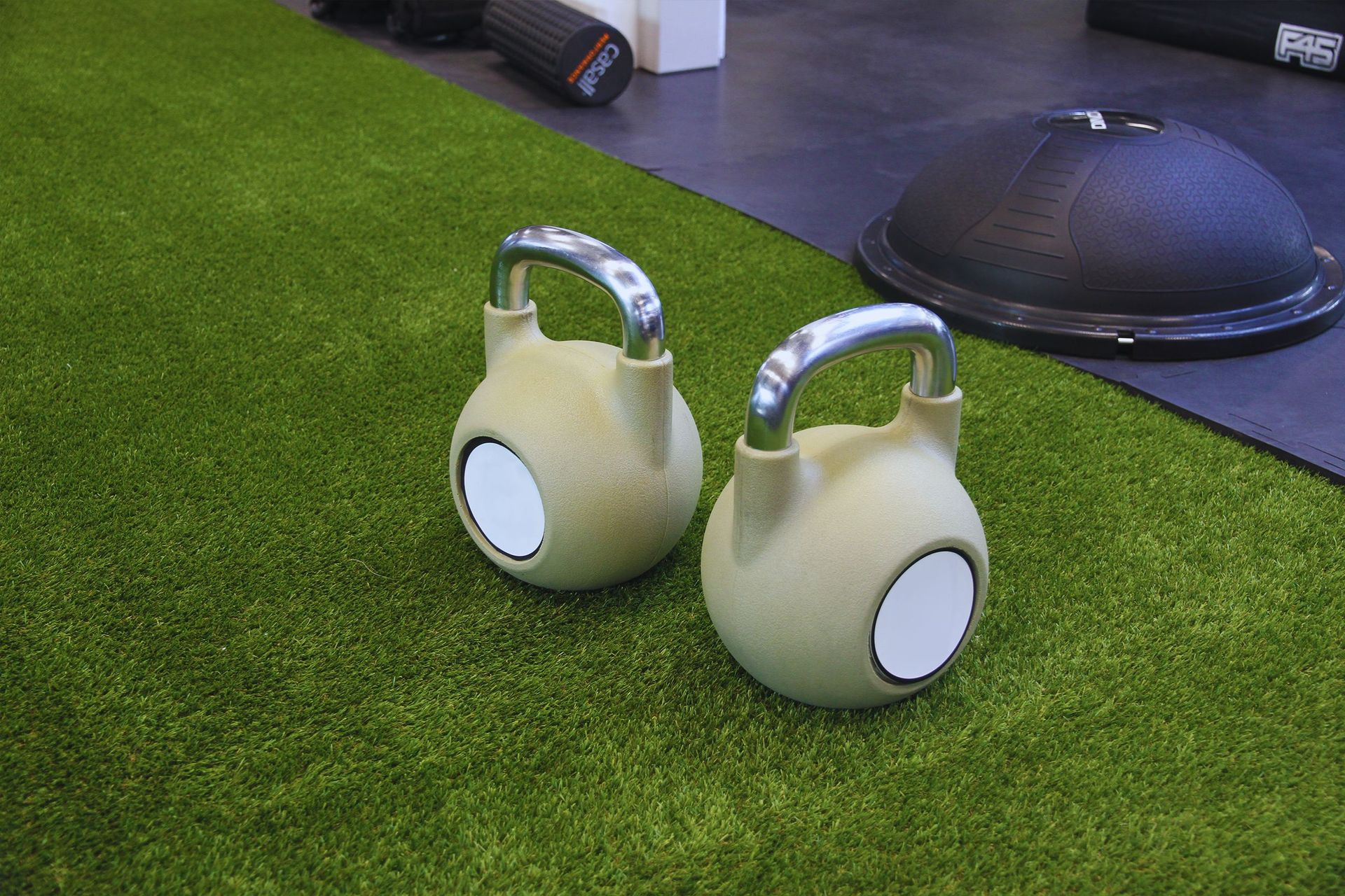 Pair of beige kettlebell-like objects on green turf near a black mat and exercise equipment.