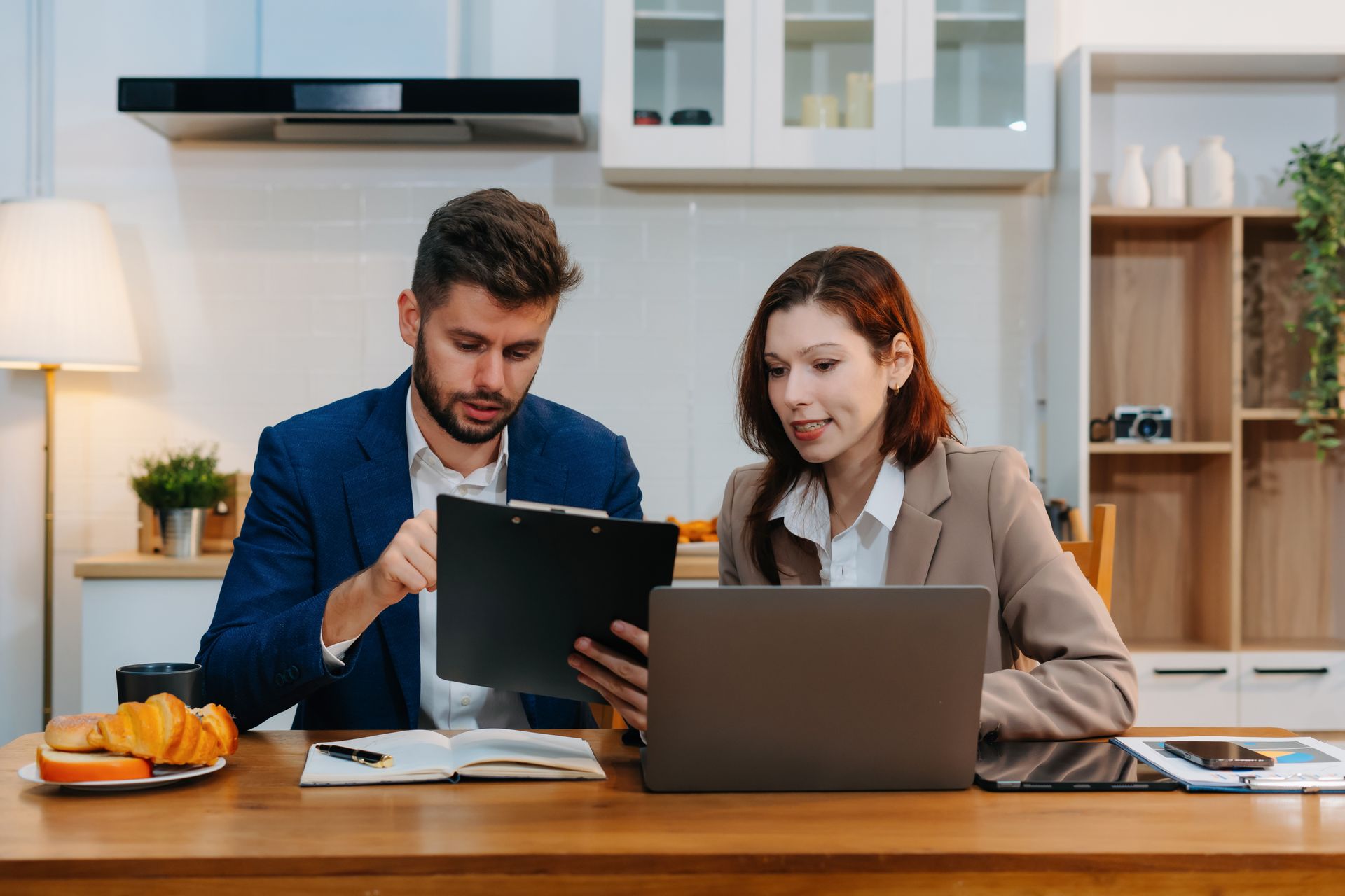 Personal financial advisor reviewing documents with client at kitchen table using laptop.