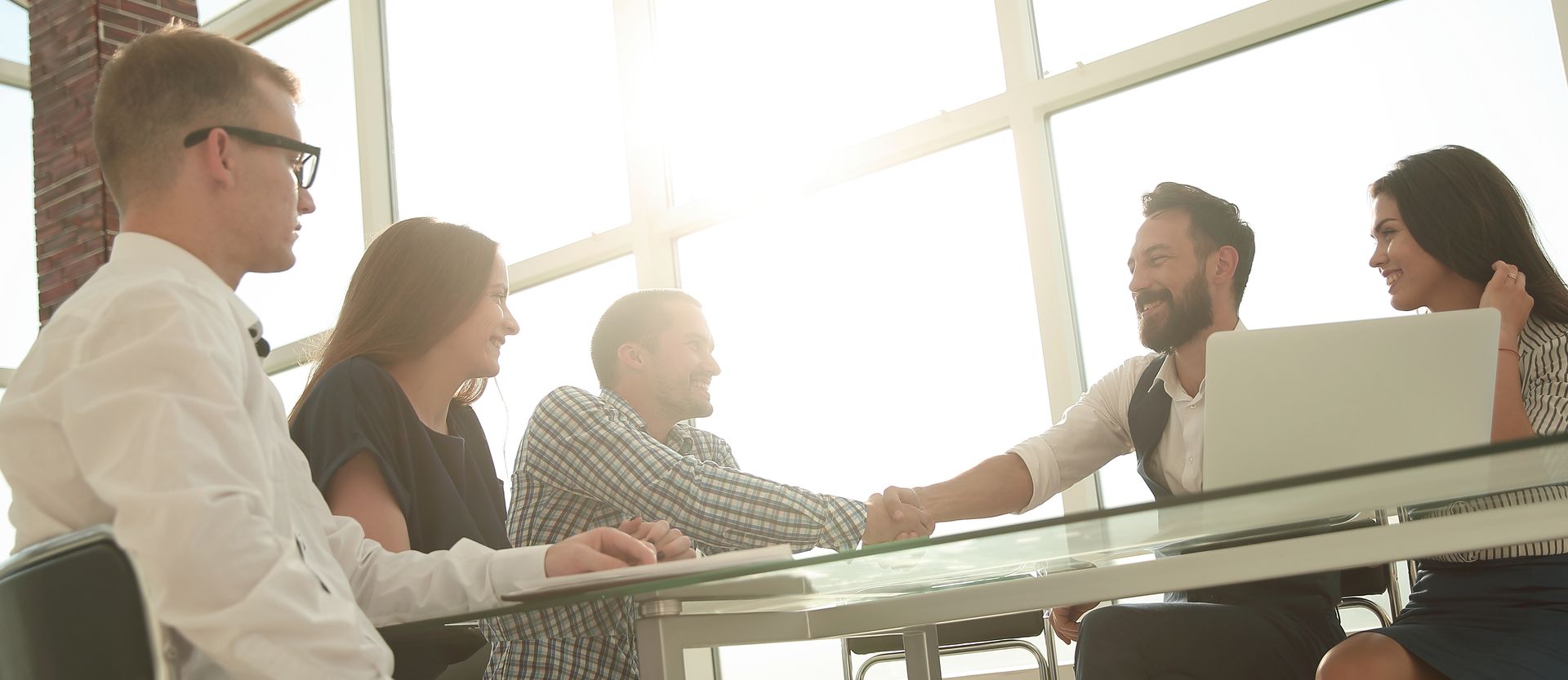 Business meeting with handshake across a desk and laptop in an office setting.