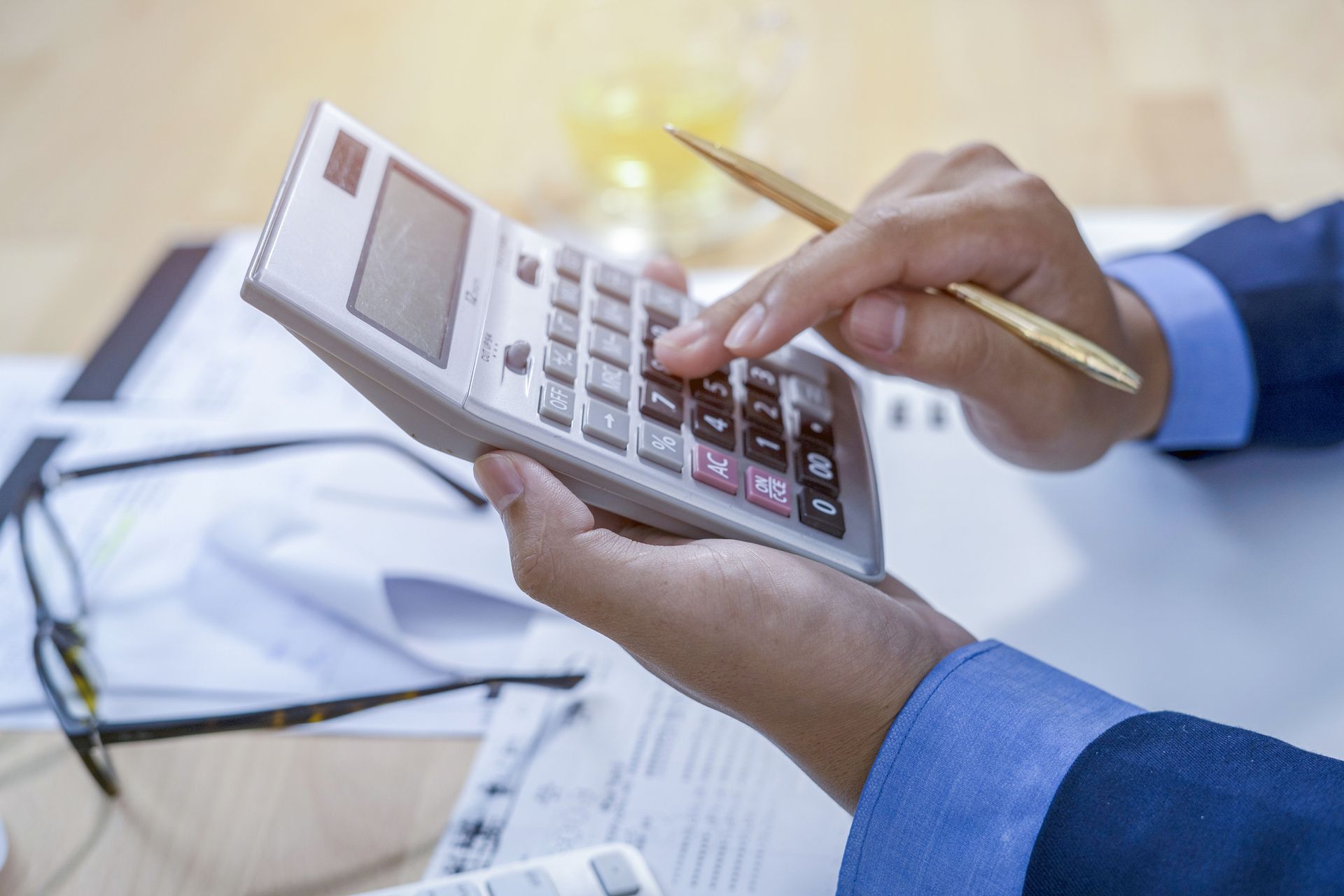 Person uses calculator with documents and glasses on desk. Person uses calculator with documents and glasses on desk.