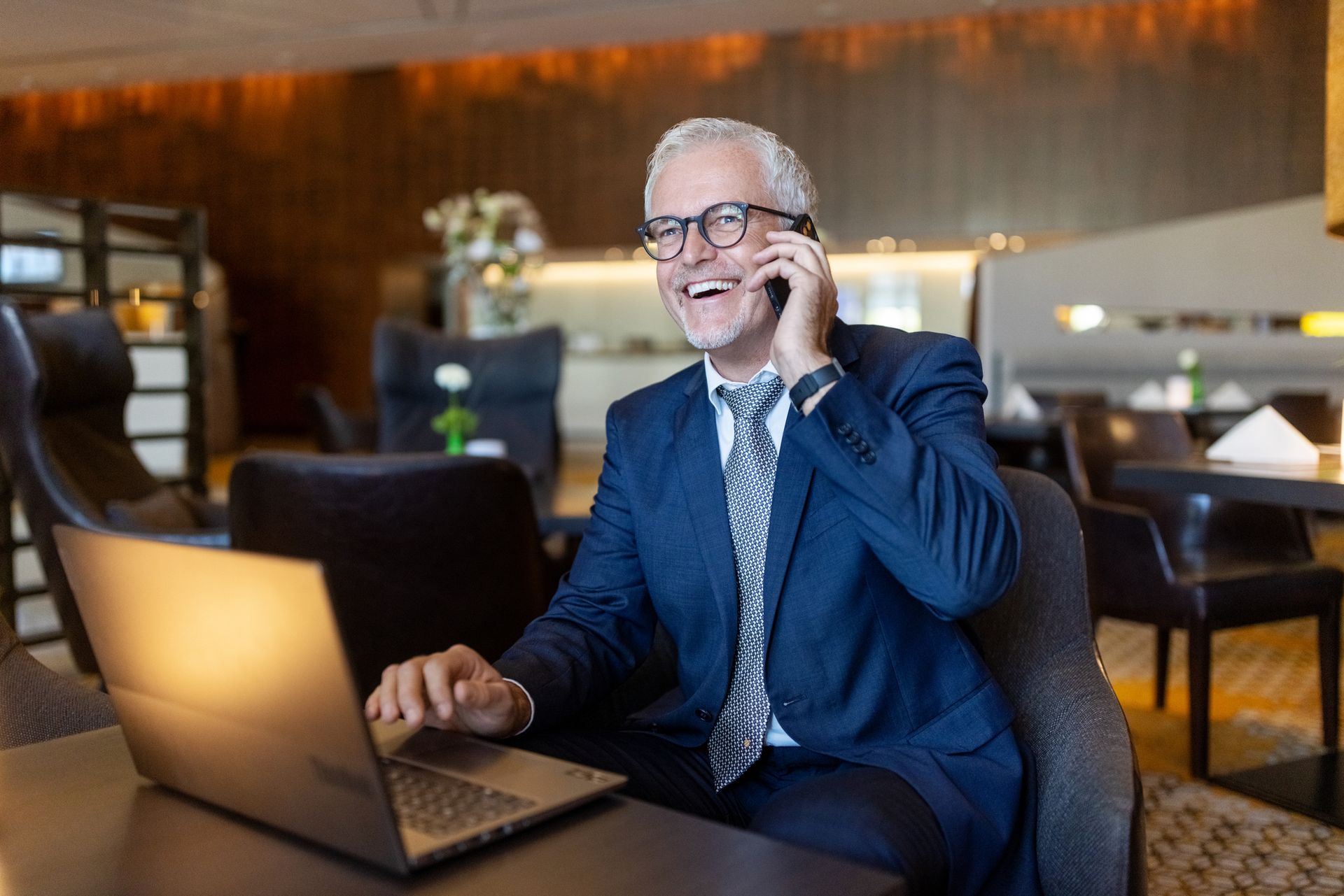 Man in suit smiles while on phone and using laptop in office.