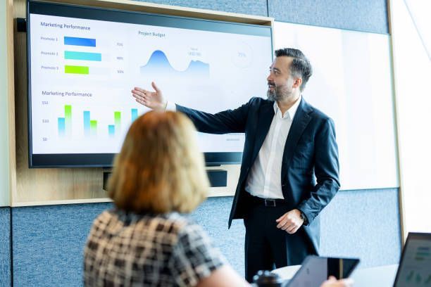A financial advisor explains a financial data report displayed on a screen in a meeting in a business office.