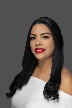 Portrait of a woman with long dark hair, wearing a white off-shoulder top and red lipstick against a gray background