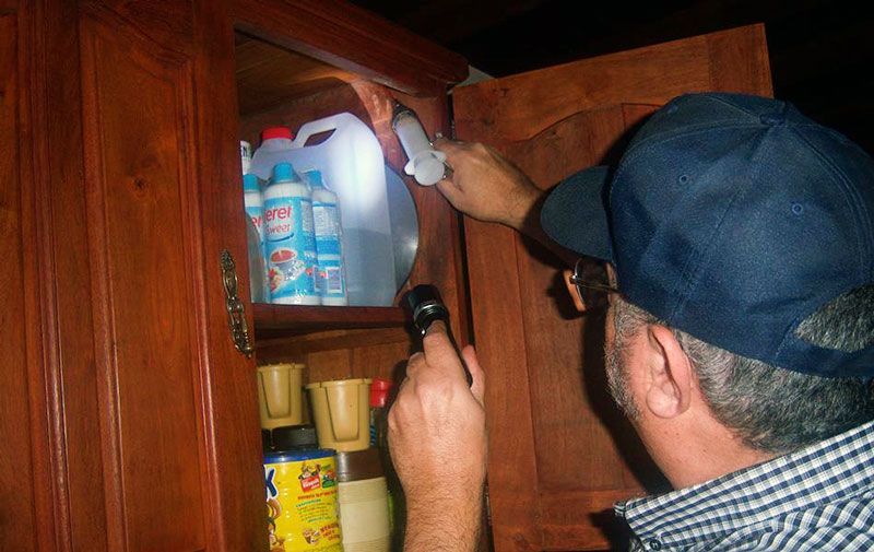 Una persona con gorra azul inspecciona un armario con una linterna; en el interior se ven productos de limpieza.