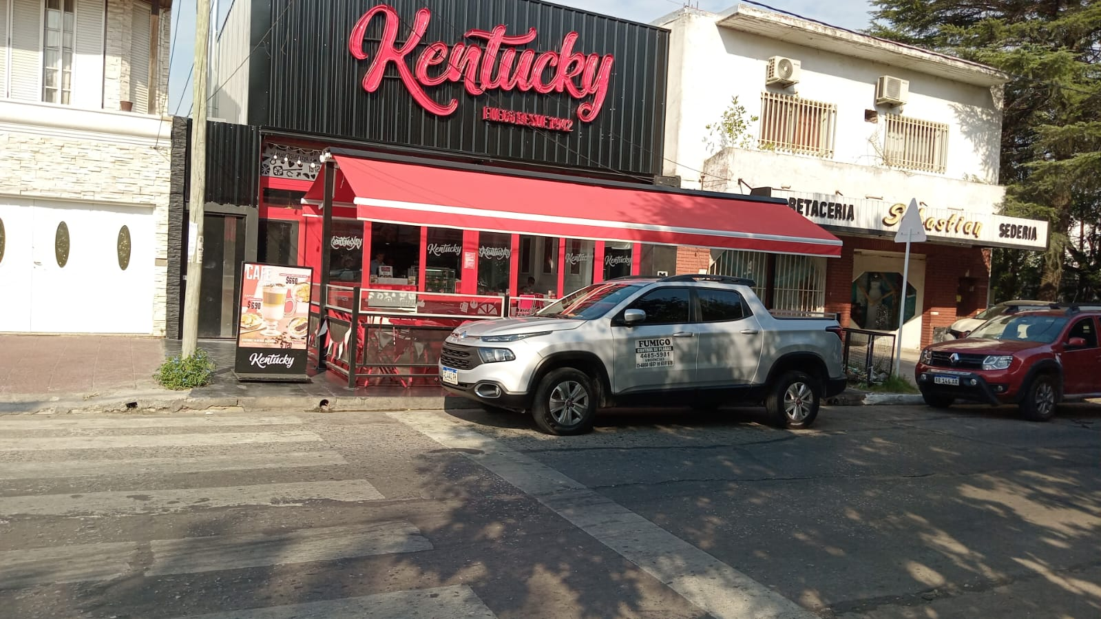Restaurante Kentucky Fried Chicken con toldo rojo, camioneta plateada y autos estacionados en la calle.