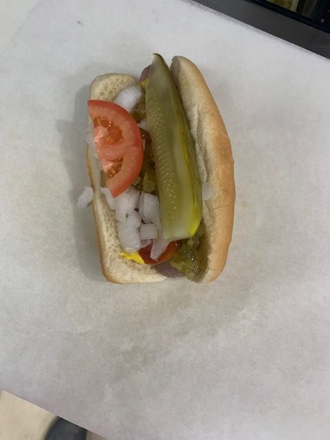 A hot dog in a bun topped with a pickle spear, diced white onions, and a tomato slice on a white surface.