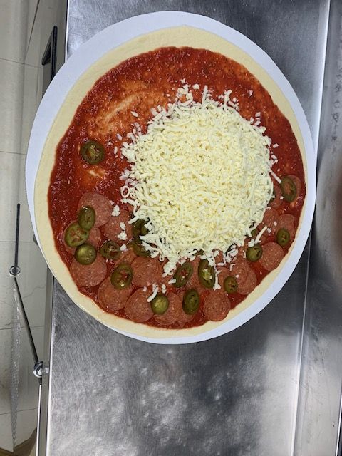 An unbaked pizza on a metal counter topped with red sauce, pepperoni slices, jalapeños, and a mound of shredded cheese.