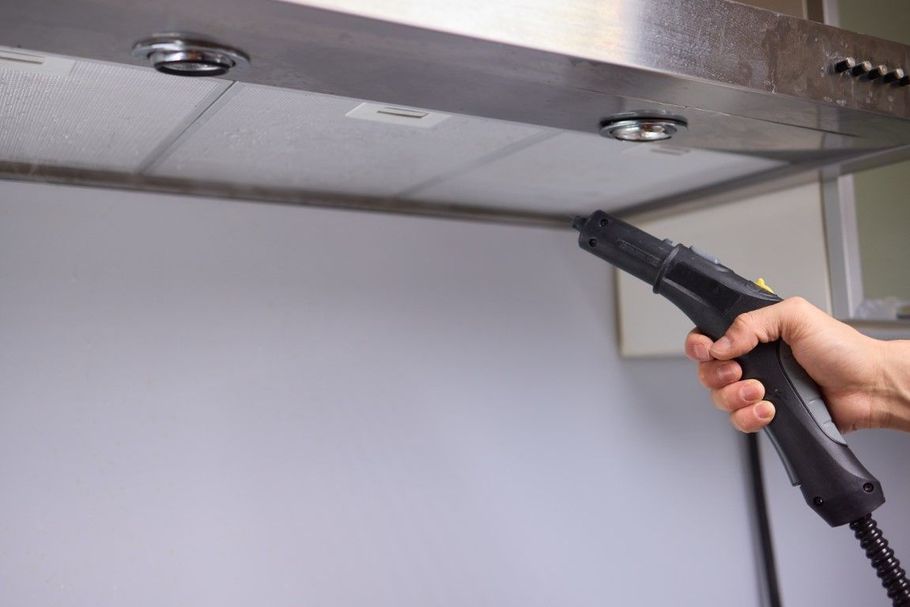 A hand uses a steam cleaner nozzle to clean the underside of a metal kitchen range hood.