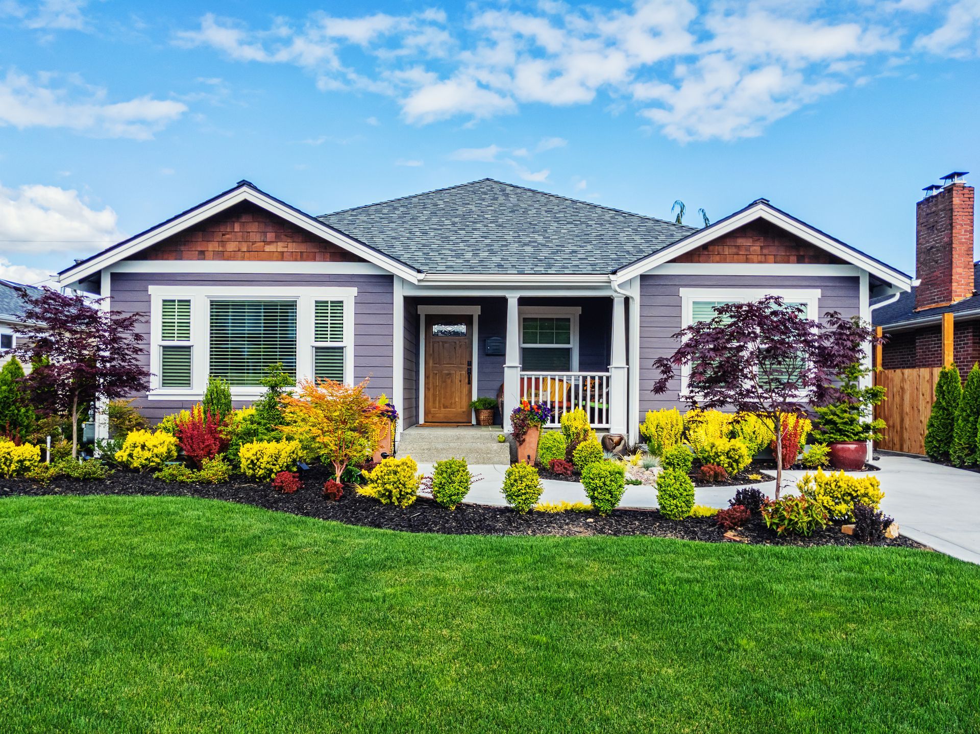 Single-story house with gray roof, front porch, colorful garden, and green lawn under a blue sky