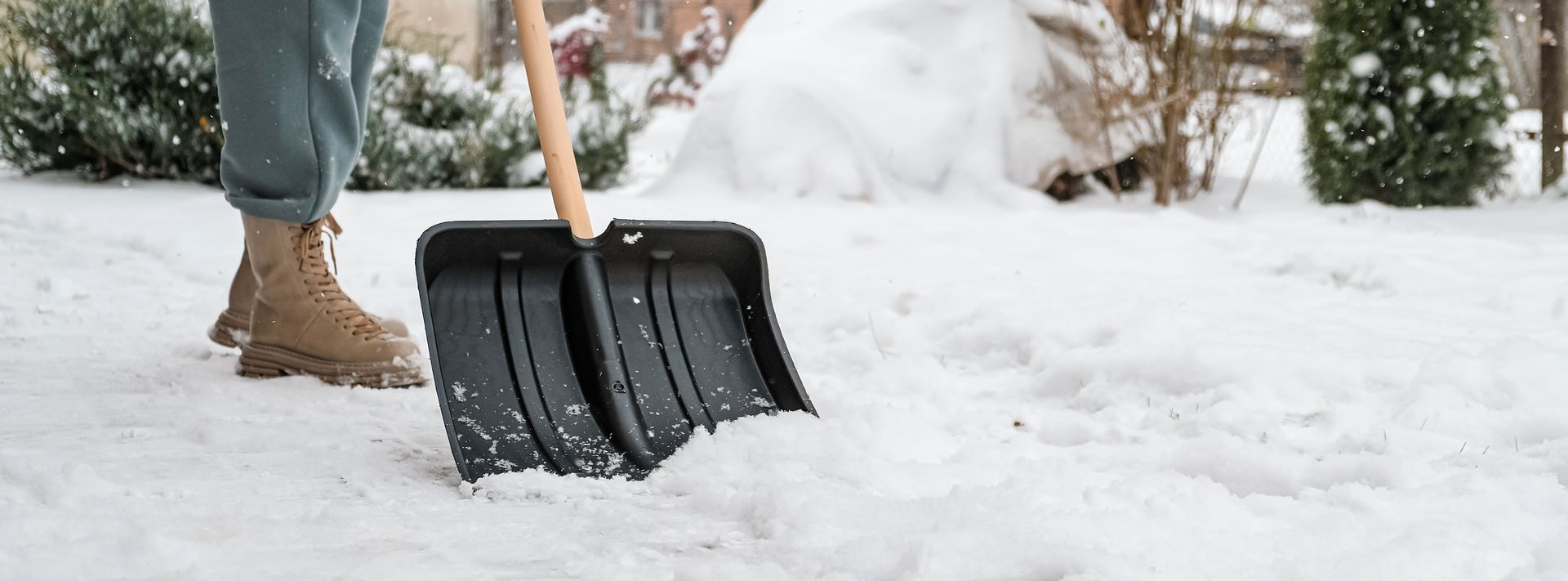 Person schaufelt Schnee auf einer schneebedeckten Fläche. Sie trägt Jeans und braune Stiefel.