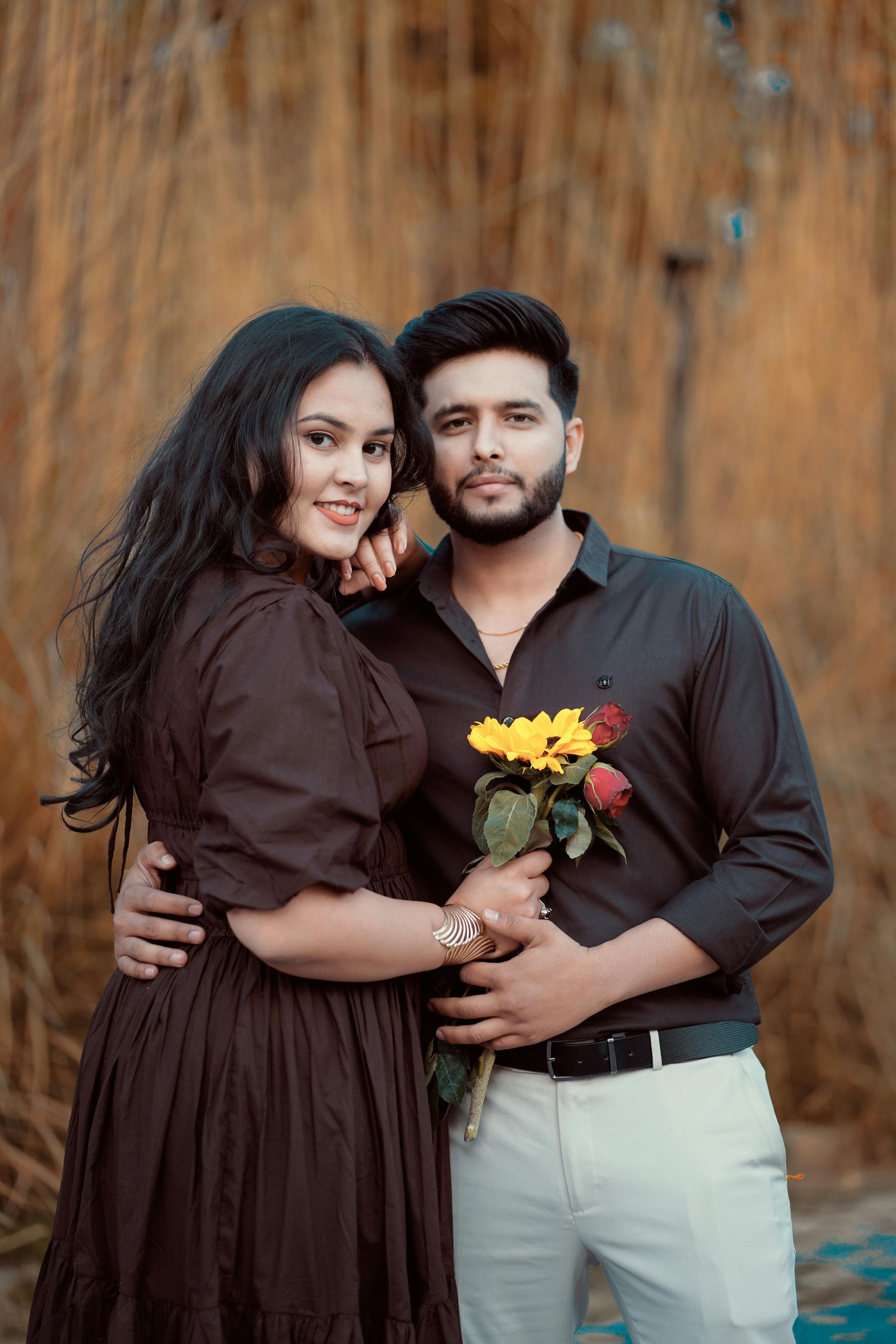 Couple standing together outdoors, smiling, with the woman in a brown dress and the man holding yellow flowers.