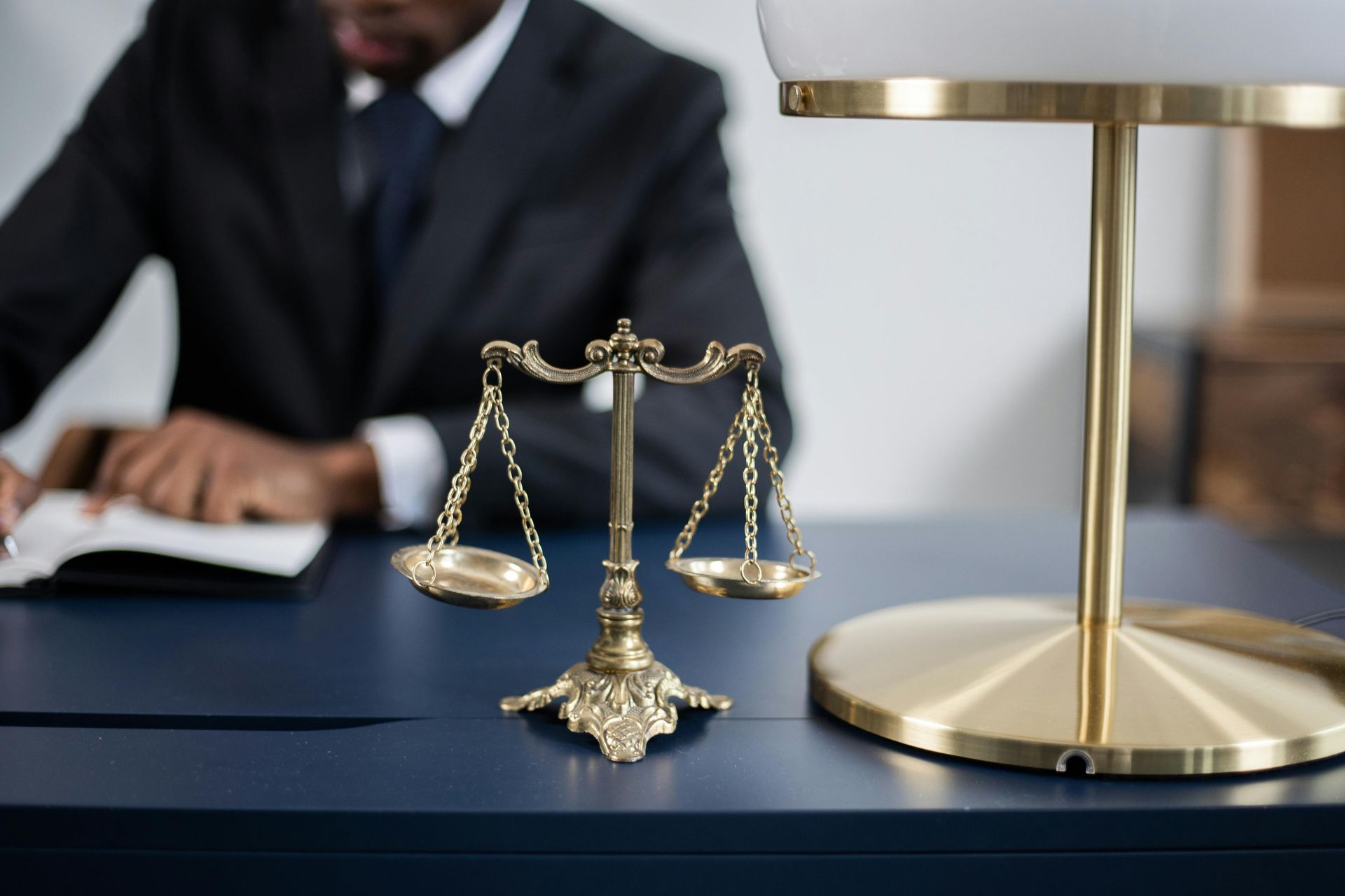 Law books and a brass scales of justice on a desk, with a blurred lawyer writing in the background