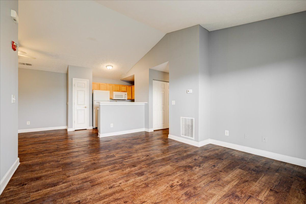 An empty living room with hardwood floors and a kitchen in the background.