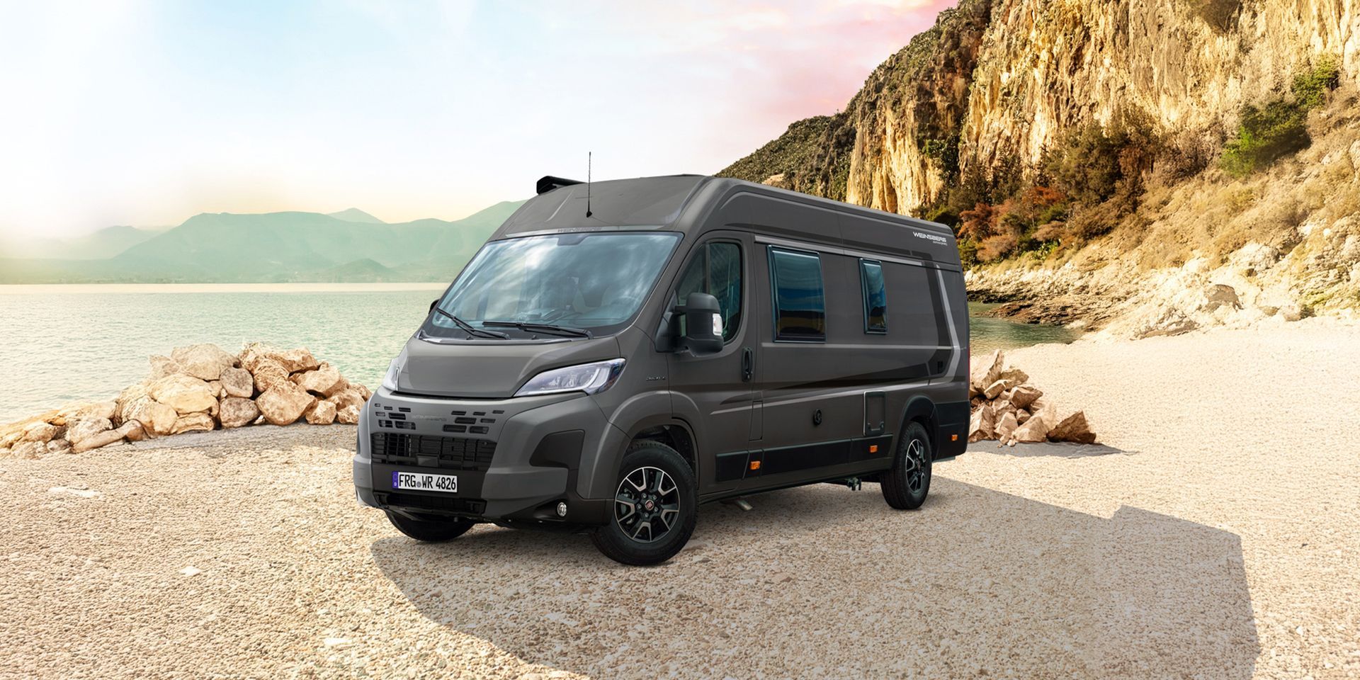 Dark gray camper van parked on a sandy beach, with mountains and water in the background.