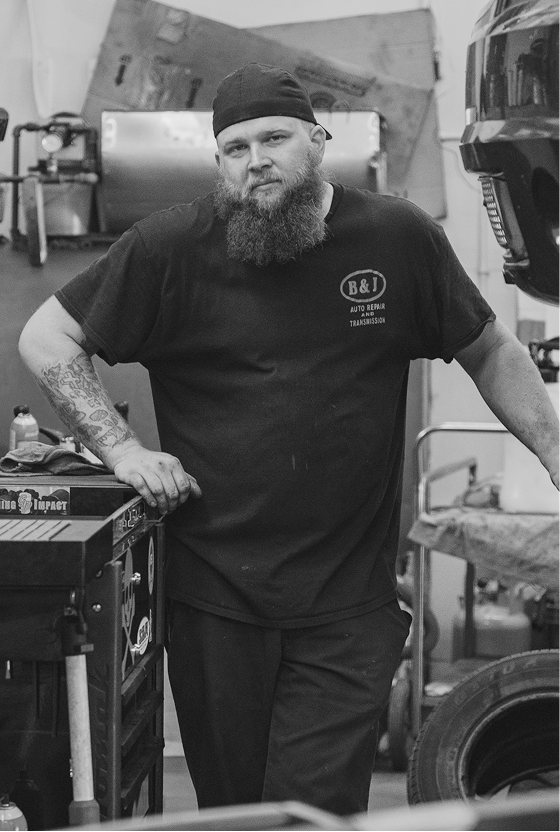 Man in mechanic shop, leaning on a toolbox, wearing a black cap and shirt with a long beard.