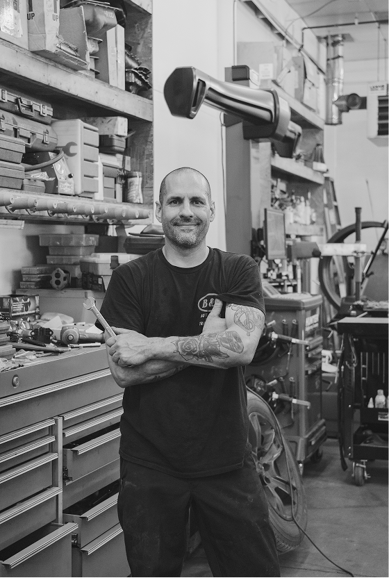 Man with tattoos smiles in workshop, arms crossed, tools and machinery visible.