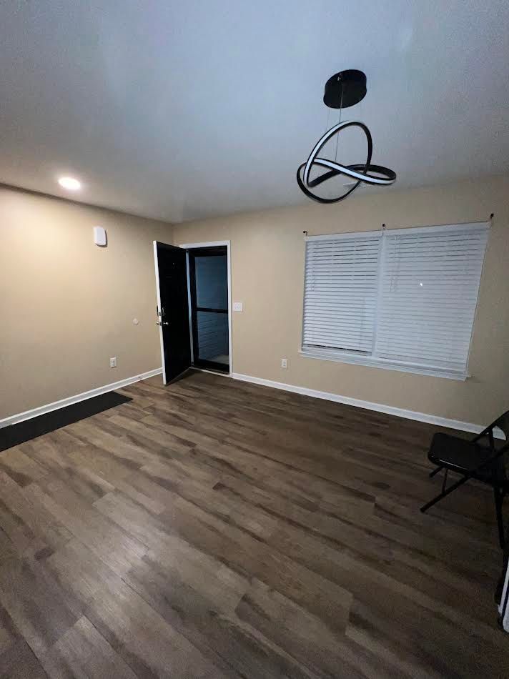 Empty room with wood floors, beige walls, ceiling light, open door, and a window with blinds.