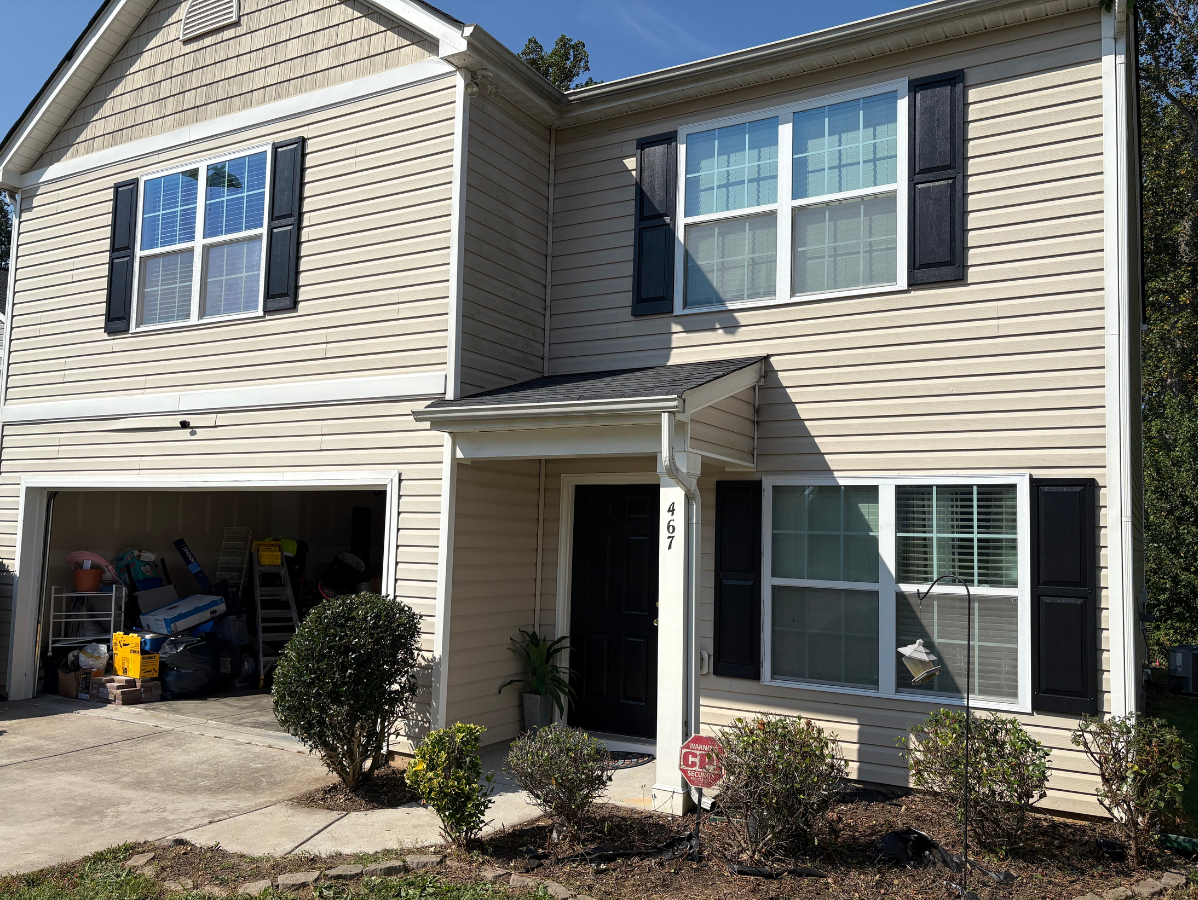 A two-story house with beige siding, black shutters, a one-car garage, and a small front yard with shrubs.