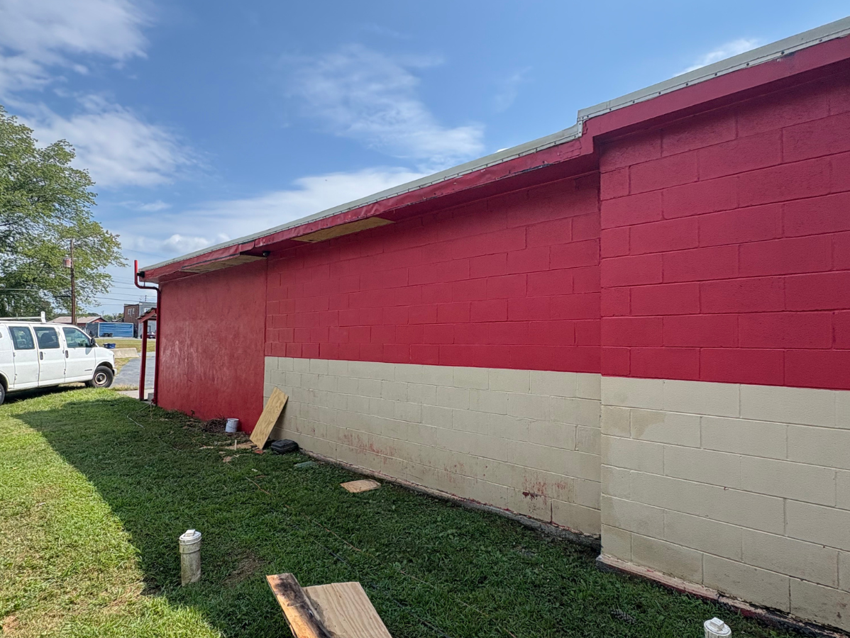 Exterior view of a red-painted concrete block wall with a tan-painted lower section, grass, and a white van in the distance.