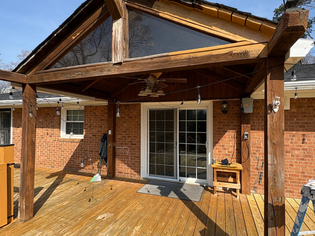 A wooden deck with a covered porch attached to a red brick house featuring a glass sliding door and a ceiling fan.