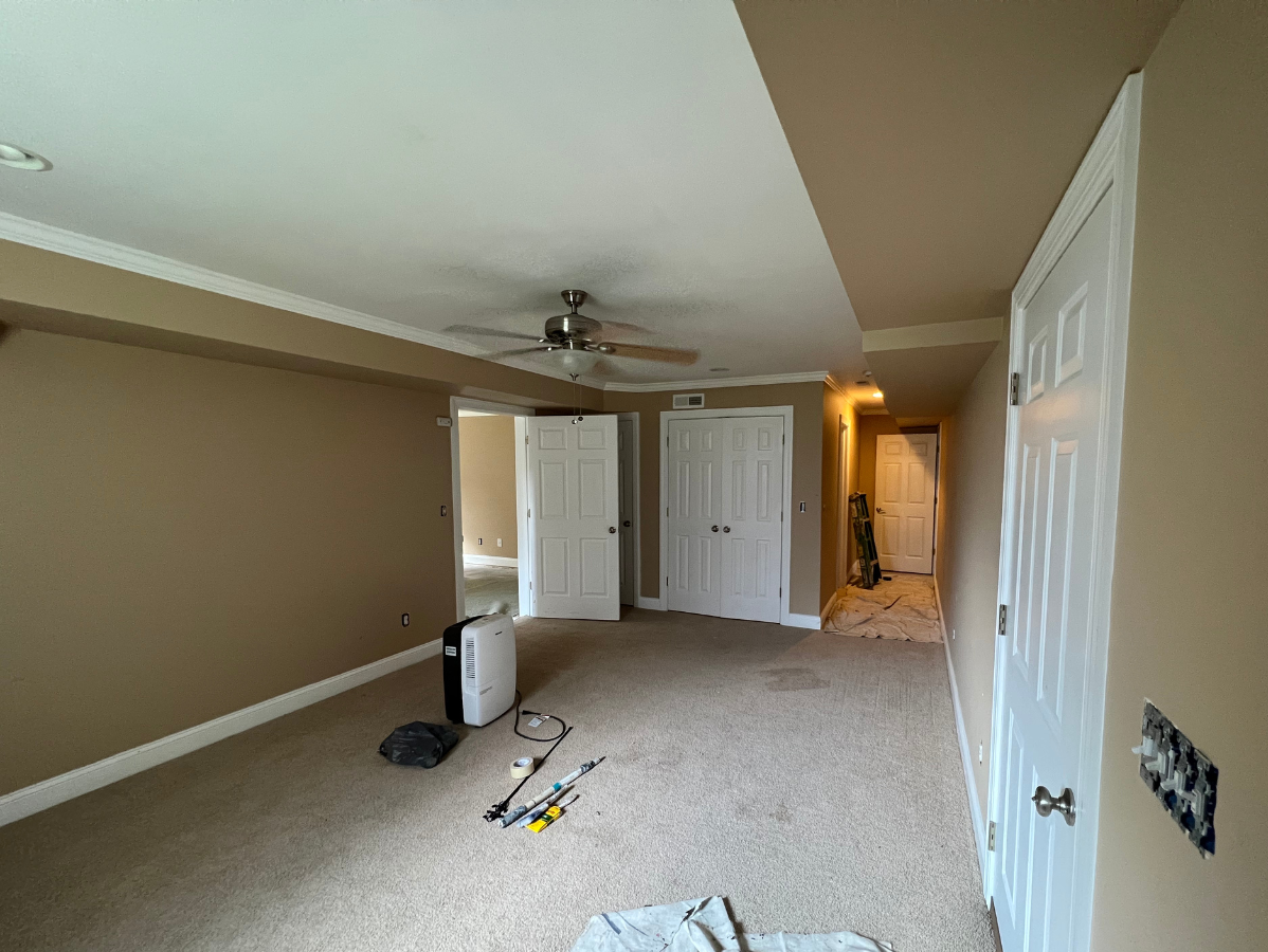 A spacious room with beige walls, tan carpet, a ceiling fan, white doors, and a portable air conditioner on the floor.