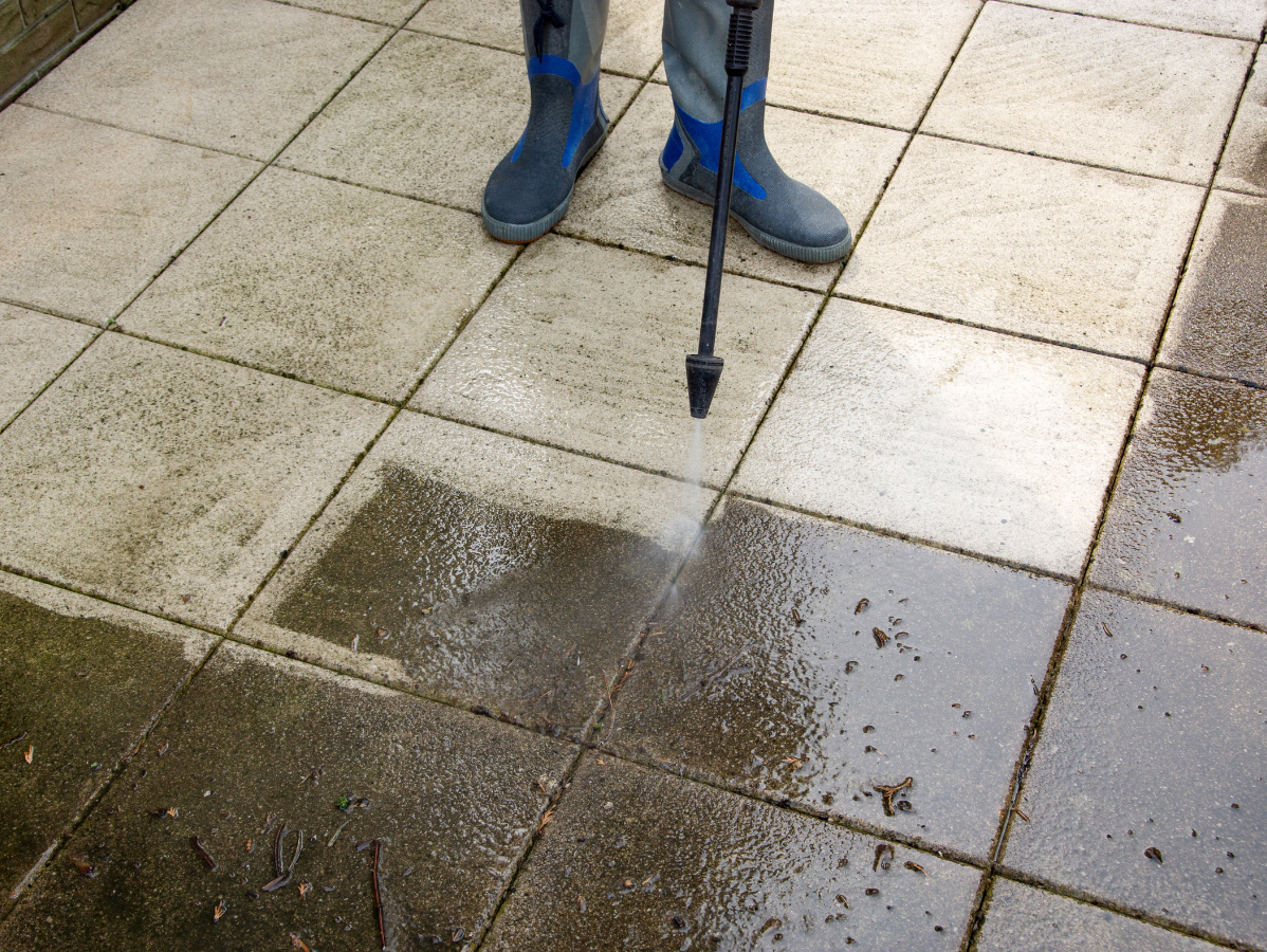 A person wearing boots uses a pressure washer to clean dirty stone paving tiles on a patio.