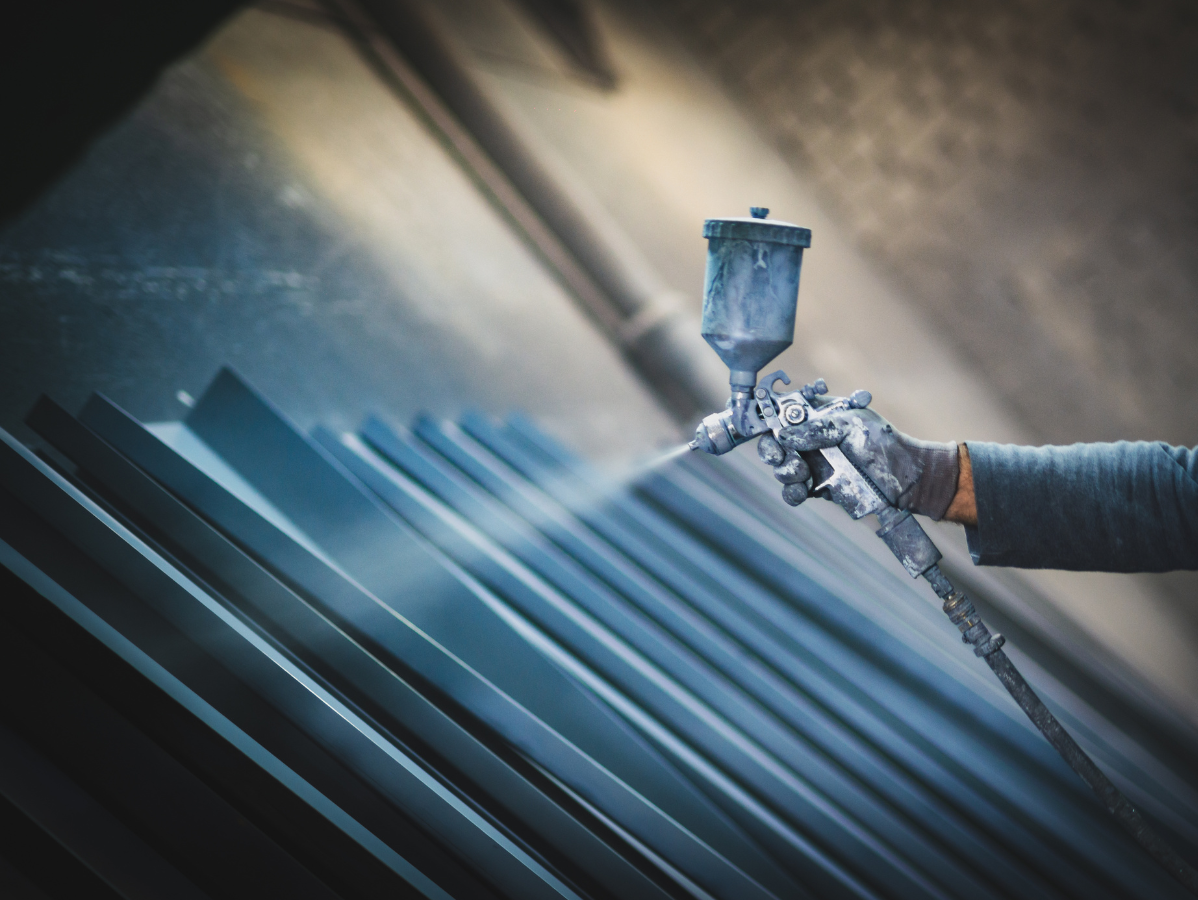 A hand holds a spray gun, applying a fine mist of blue paint onto a series of dark metal slats.
