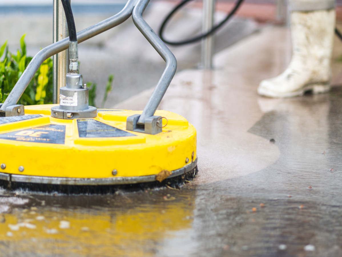 A yellow surface cleaner power-washing a wet concrete sidewalk, with a person in white boots visible in the background.
