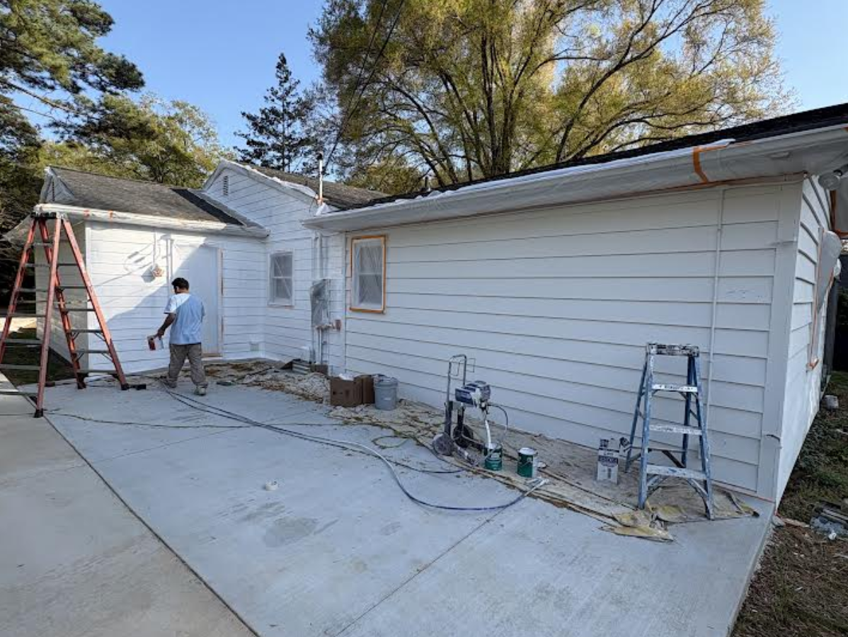 Worker on a ladder beside a white house, near a garage in a driveway during exterior repairs.