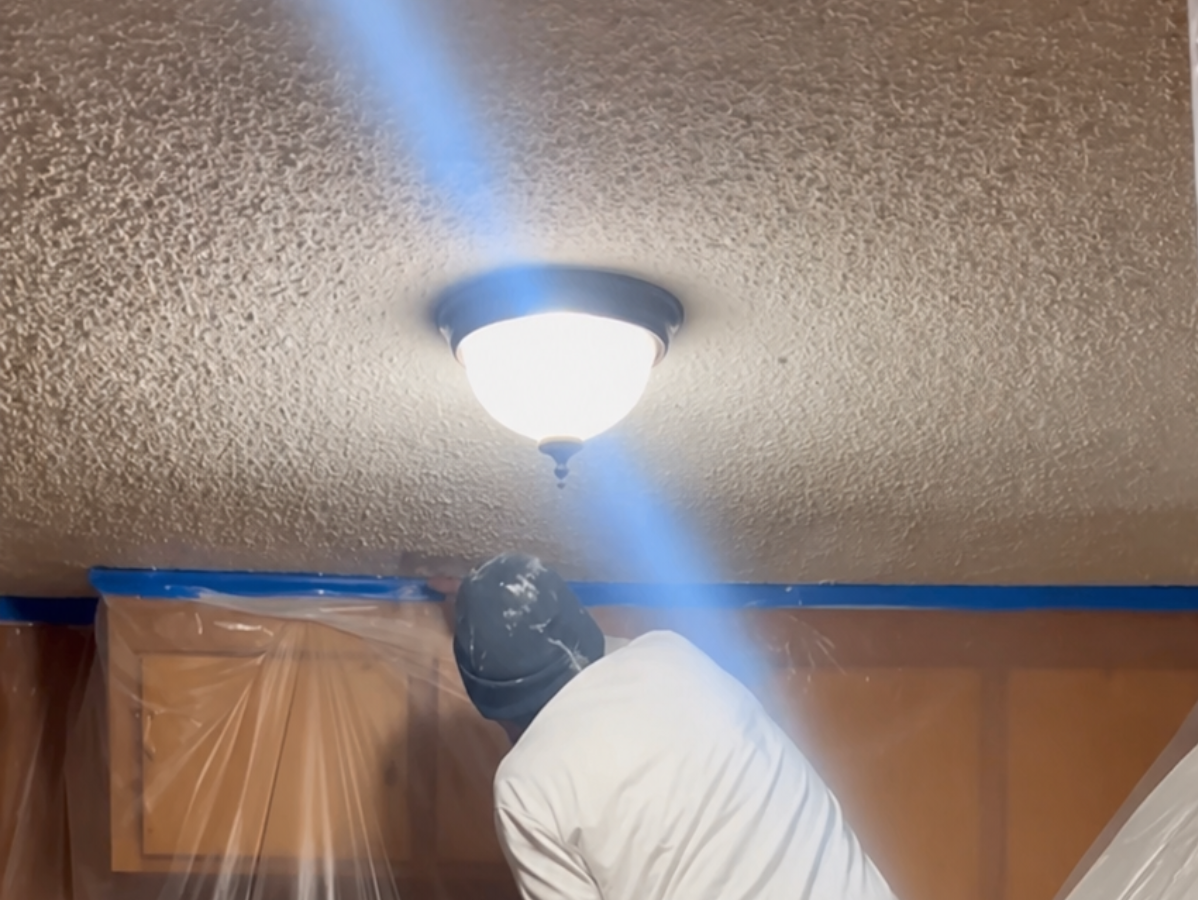 Person in white hoodie painting a ceiling light fixture in a room, with blue painter’s tape along the walls.